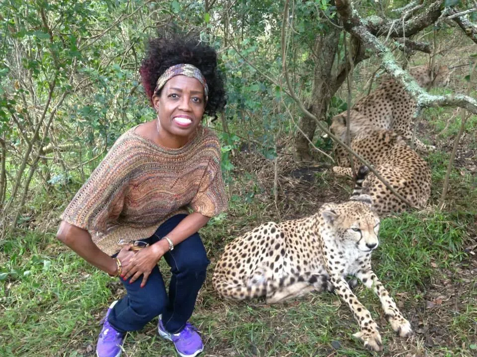 Coach Judy Handwerker kneels beside a resting cheetah in a natural wildlife setting surrounded by trees and greenery. Smiling toward the camera, she wears casual outdoor attire and purple athletic shoes while posing near the majestic animal. The image highlights adventure, courage, connection with nature, travel experiences, and an active, vibrant lifestyle.