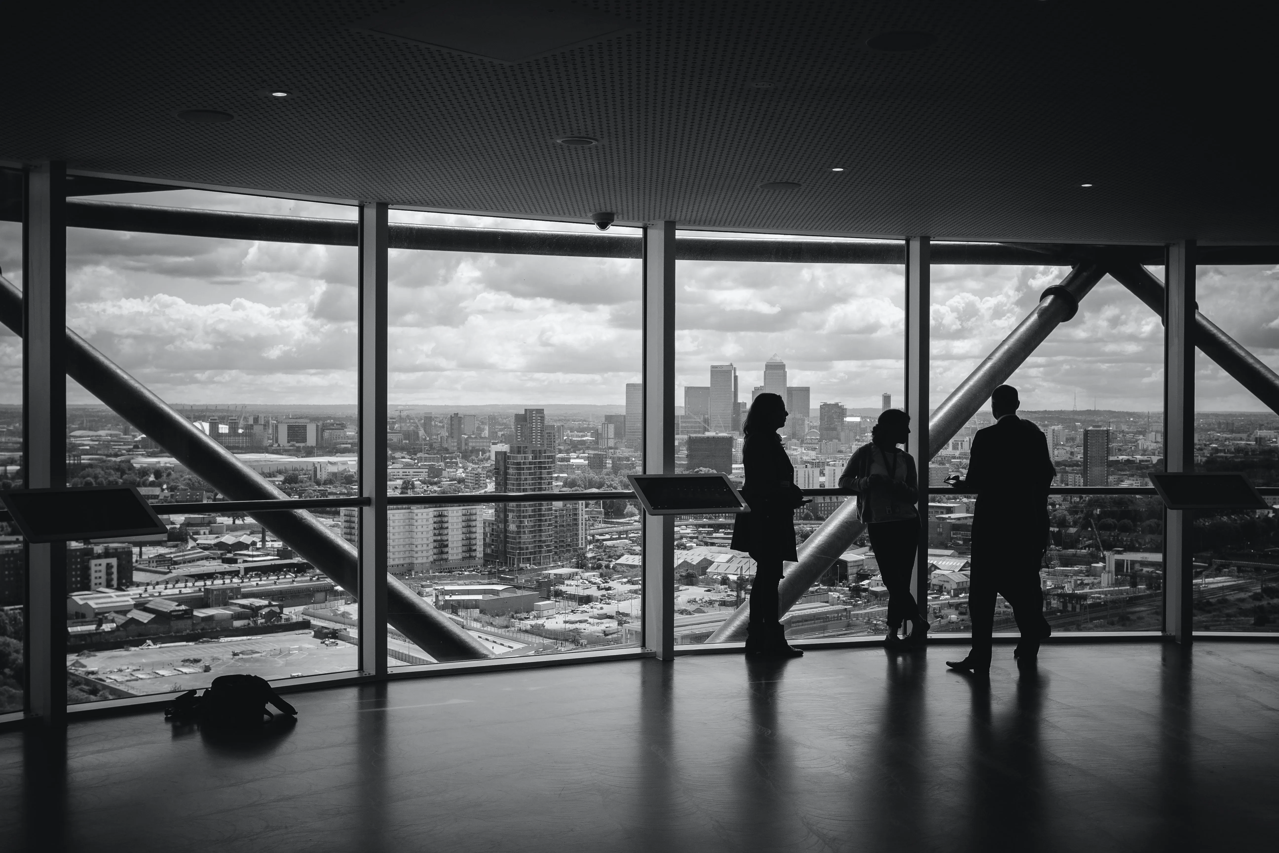 Black-and-white photo of three people standing in front of large windows overlooking a city skyline. The individuals appear to be engaged in conversation, silhouetted against the urban backdrop of skyscrapers and cloudy skies.