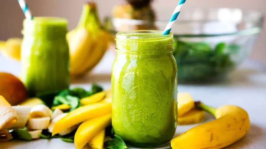 Two mason jars of green smoothies surrounded by fruits like bananas and berries.
