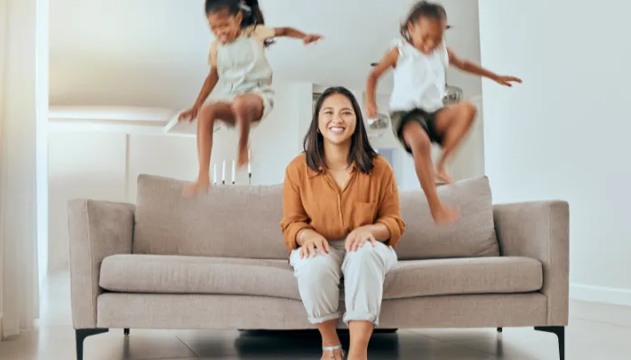 A mom on a couch smiling while her kids are jumping on it.