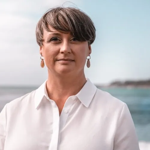 A closeup headshot image of Director-Founder, Michele Hurlburt. She is facing the camera wearing a white, button up blouse with the ocean behind her.