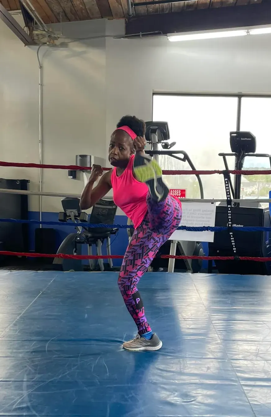 Coach Judy Handwerker demonstrates strength, flexibility, and athletic movement inside a boxing gym, performing a high kick while balancing on one leg in vibrant pink fitness attire. Surrounded by a boxing ring and exercise equipment, she showcases functional fitness, mobility, balance, and active aging for women over 40.