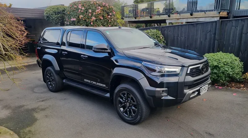 A black Toyota Hilux SR5 Cruiser pickup truck is parked in a driveway near a modern home with a black wooden fence. The vehicle features black alloy wheels, tinted windows, and a canopy over the truck bed. Surrounding the driveway are green bushes, flowering plants, and a tree with bare branches.