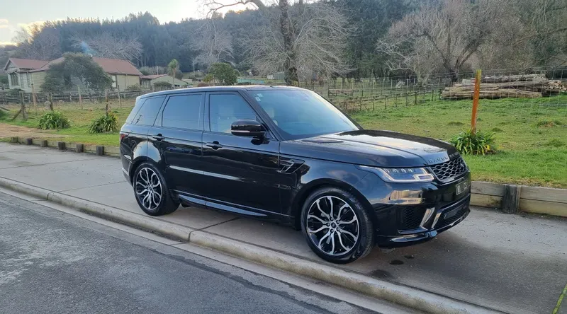 A black Range Rover Sport is parked on the side of a suburban road in a rural area. The SUV features black and silver alloy wheels, tinted windows, and a sleek, glossy finish. The background includes green fields, trees, and a few houses, with a partially cloudy sky and a serene, late afternoon atmosphere.