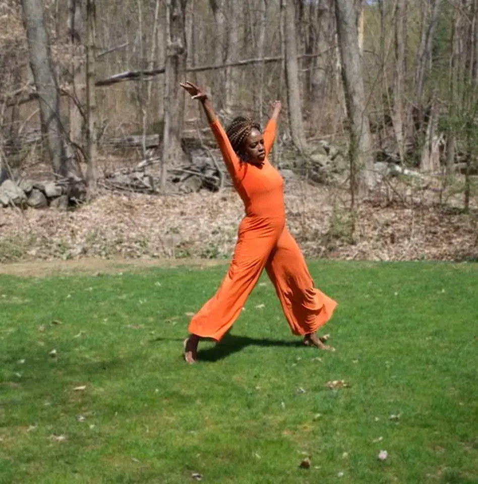 Coach Judy Handwerker performs an expressive movement pose outdoors on a grassy lawn, wearing a flowing orange outfit with arms raised gracefully overhead. Surrounded by trees and natural scenery, she demonstrates balance, flexibility, joyful movement, and whole-person wellness in a peaceful outdoor setting.
