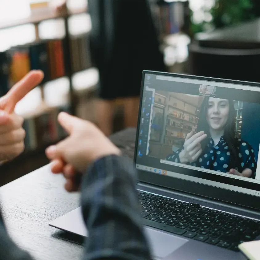 A modern laptop on a table shows a virtual meeting with two people using sign language. One person, wearing a polka-dotted shirt, is animatedly signing. In the foreground, a hand is raised, indicating active participation. Behind the laptop, soft shelving suggests a studious environment. A banner at the top reads 'SPEAKING,' emphasizing communication skills.