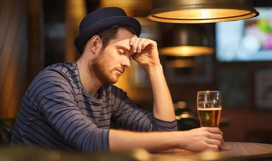 a man sitting alone at a bar, resting his head on one hand while holding a glass of beer with the other. He appears to be deep in thought or possibly feeling down or stressed. 