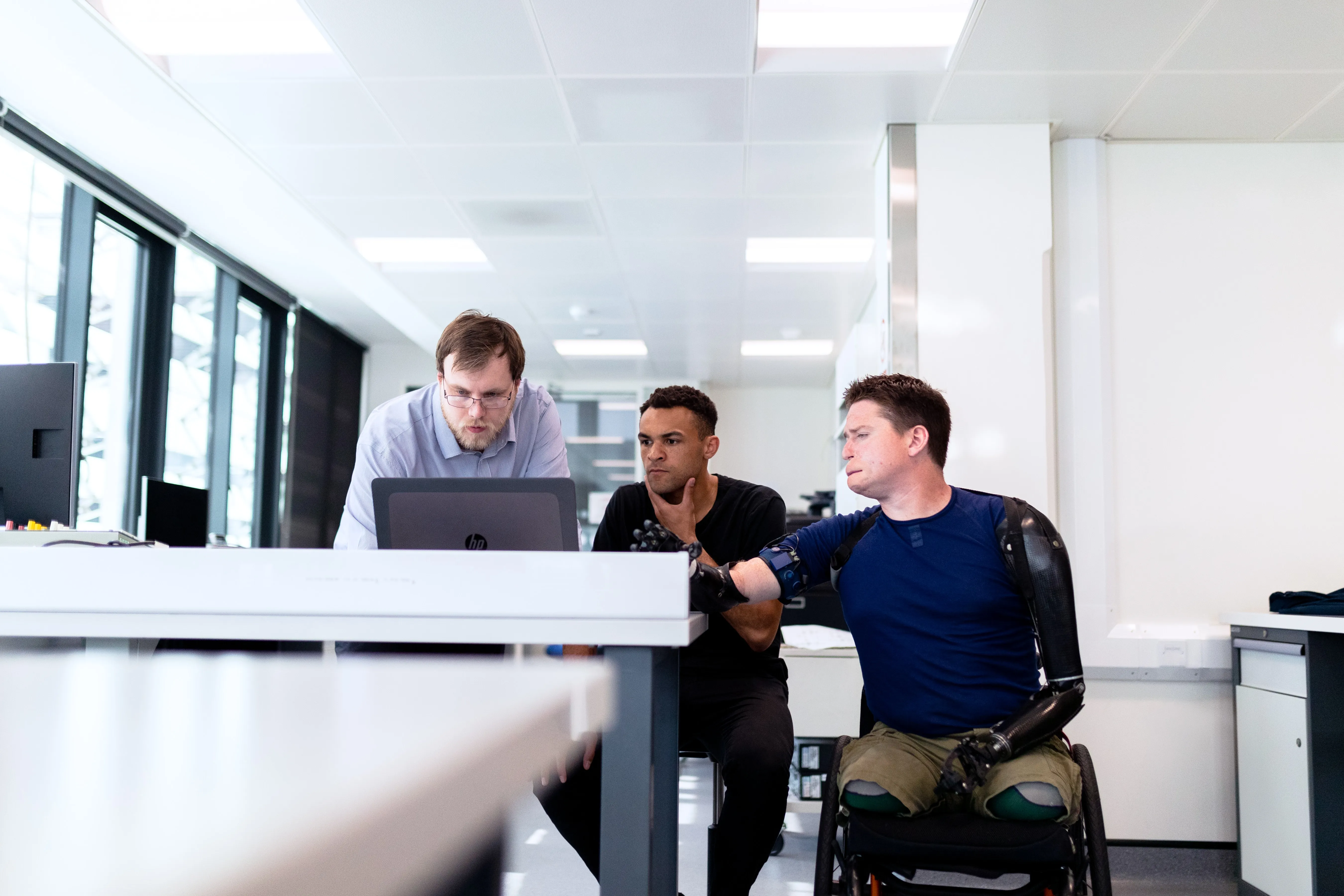3 male employees sitting and working together.  One employee is sitting in his wheelchair with his prostetic legs and left arm