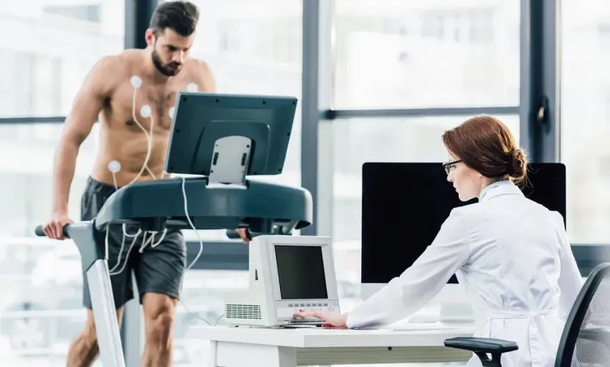 a woman in a lab coat in front of a computer running tests on a man nearby on a treadmill hooked up to monitors.