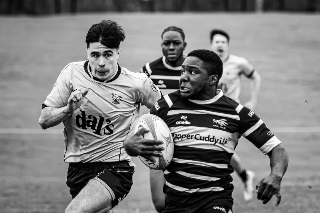 Black and white action rugby photograph showing a scared ball carrier running from a tackler