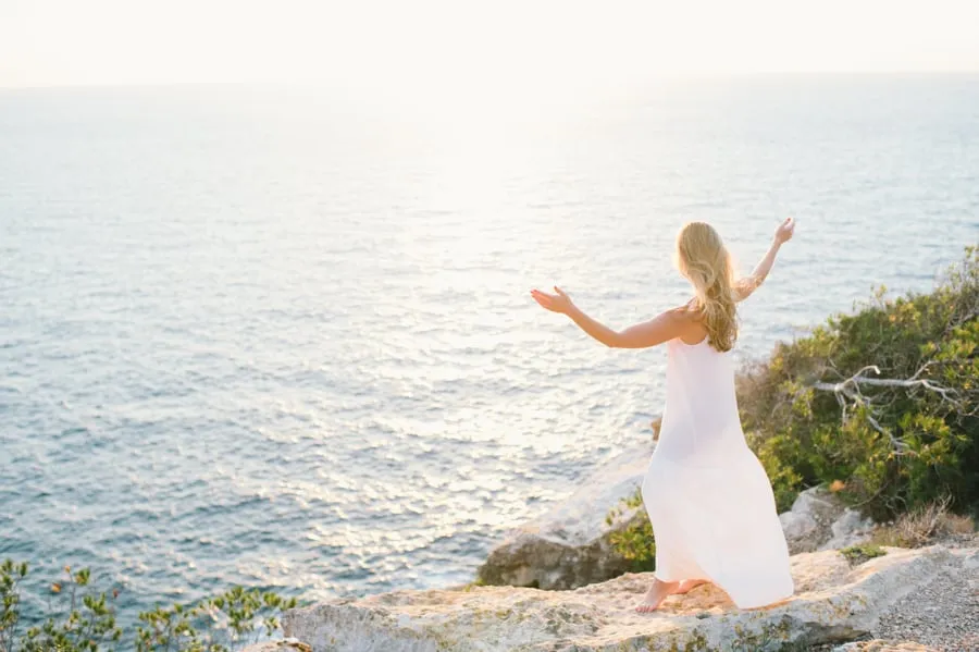 A woman standing on a rock on top of a cliff . Her arms are raised and spread out in the air. 