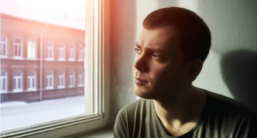  A thoughtful person sitting by a window, looking out with a subdued expression, symbolic of contemplation or melancholy.