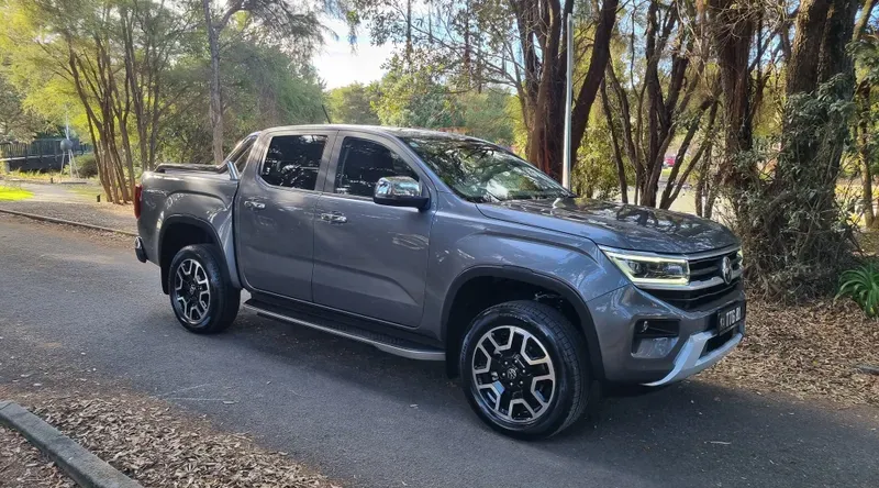 A grey Volkswagen Amarok Aventura V6 pickup truck is parked on a tree-lined road. The vehicle features black and silver alloy wheels, chrome accents, and tinted windows. The surrounding area is shaded by tall trees, with sunlight filtering through the leaves, creating a serene, natural setting. Fallen leaves cover parts of the road and curb, adding to the rustic atmosphere.