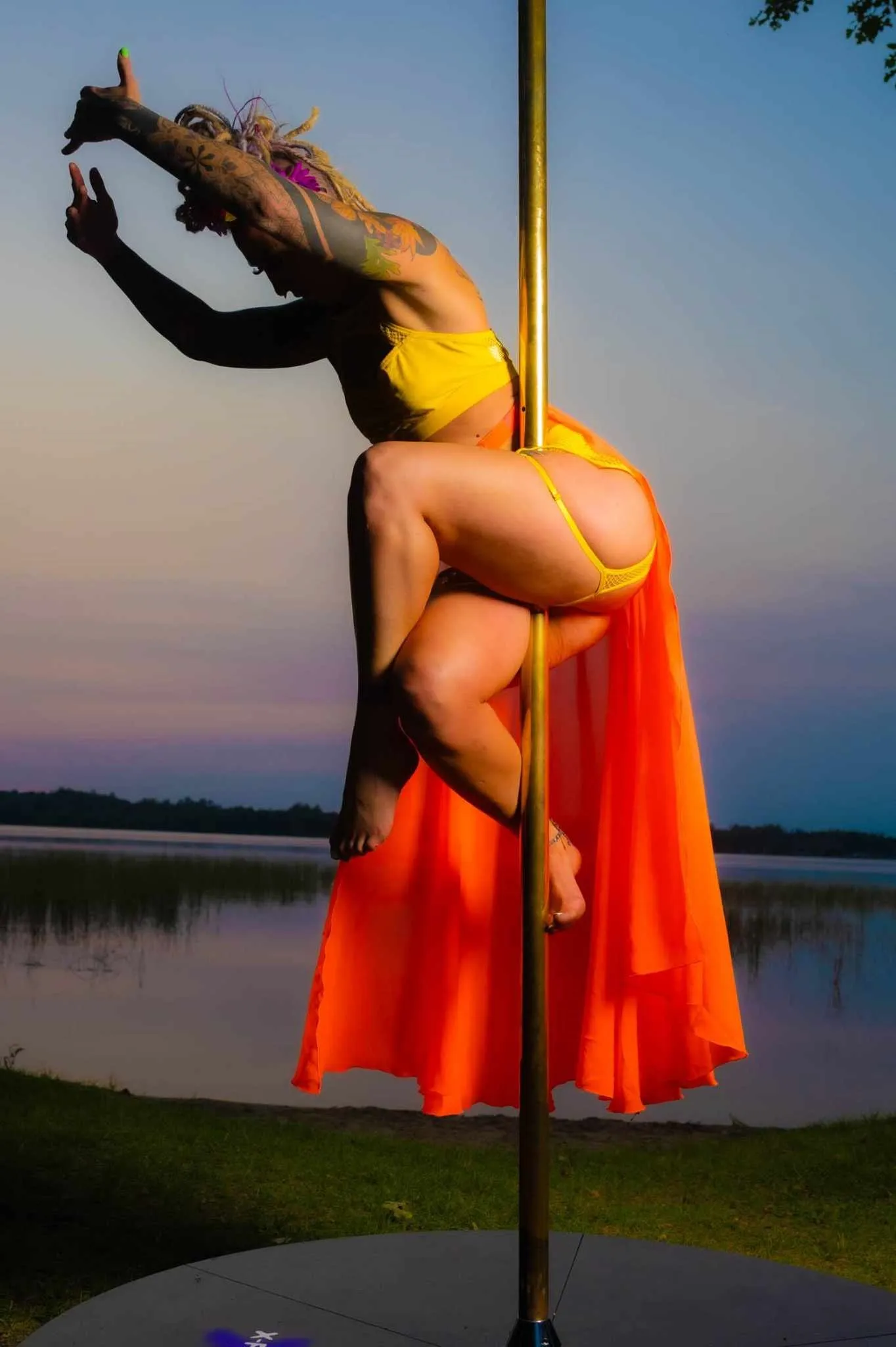 Amanda is posed in a yellow top and orange skirt in a sit on the pole with a sunset and lake in the background. 