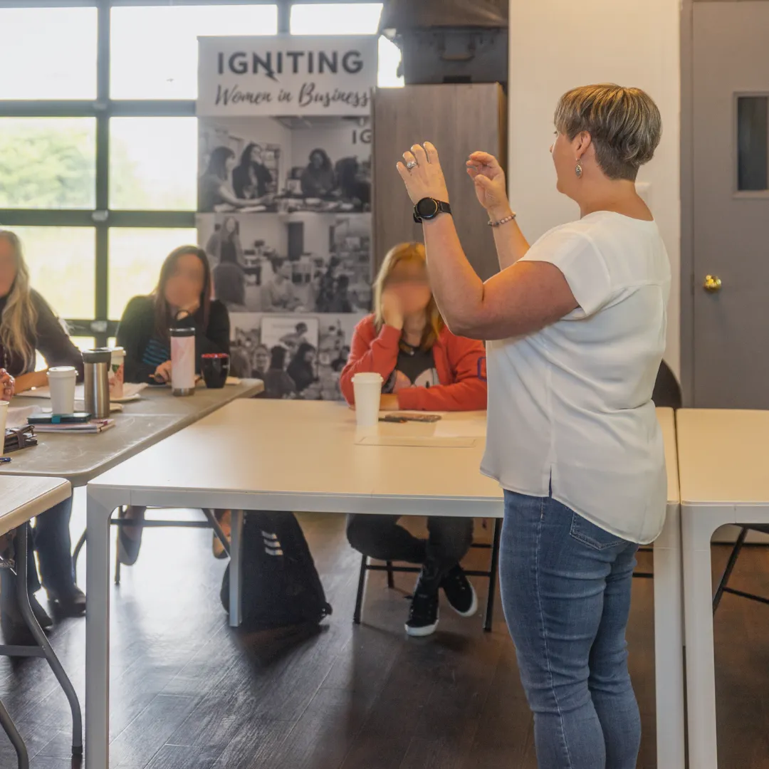 Founder, Michelle Hurlburt standing in front of folding tables and a few people, speaking with her arms raised. She is wearing jeans and a white blouse.