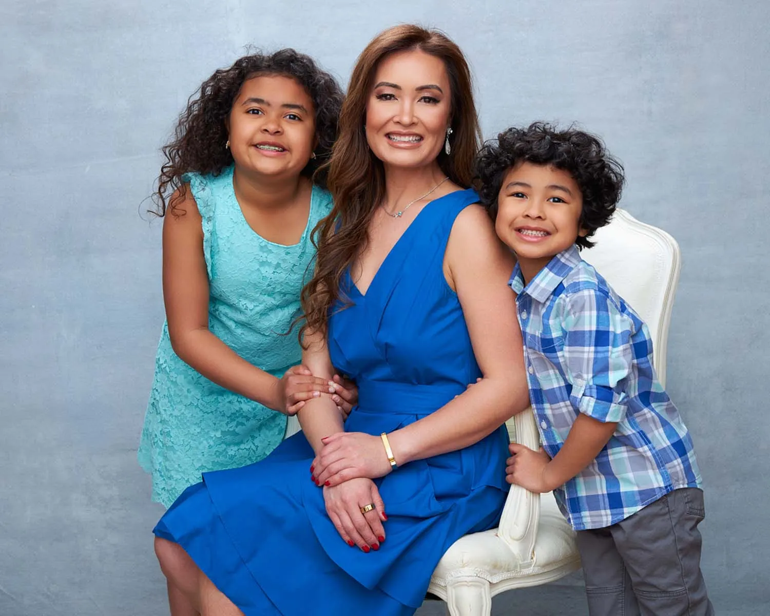 Portrait of a family of three with a mother, daughter and son all wearing shades of blue smiling and posed in front of a light blue backdrop