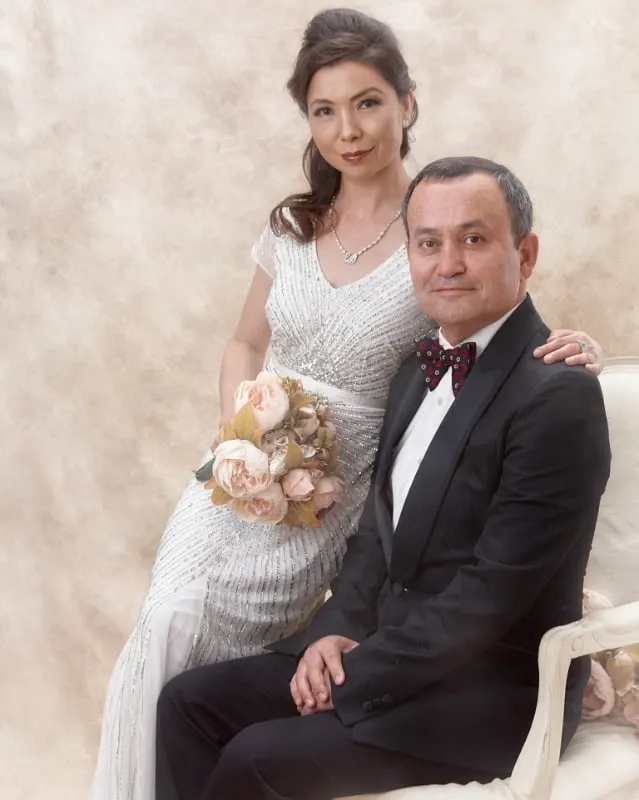 Brightly lit couples bridal portrait of a couple where the husband is in a full suit sitting and the wife is wearing a wedding dress leaning against him