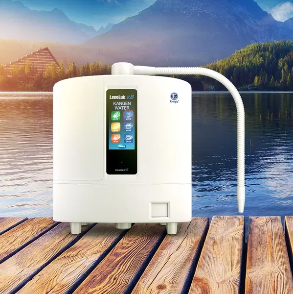 An alkaline water ionizer machine, the Leveluk K8 Kangen Water, is displayed on a wooden dock overlooking a serene lake with mountains in the background. This setting promotes the machine's purpose of transforming tap water into clean and healthy drinking water, harmoniously linked with the concept of natural purity and wellness.