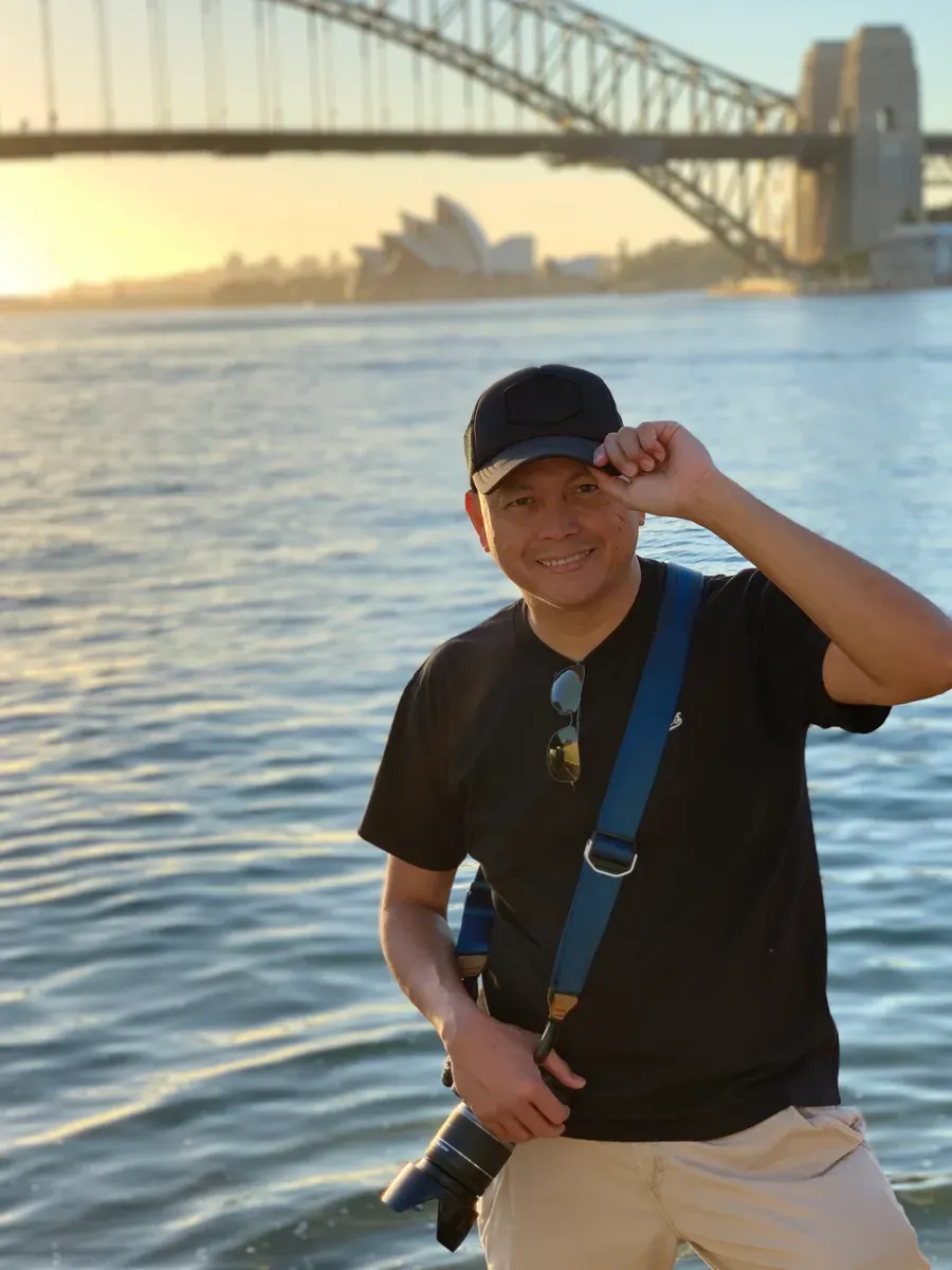 man wearing black cap, black shirt, cream pants, with camera strapped across his body, with the Sydney Harbour Bridge and Sydney Opera House visible on the background as the sun rises across the harbour