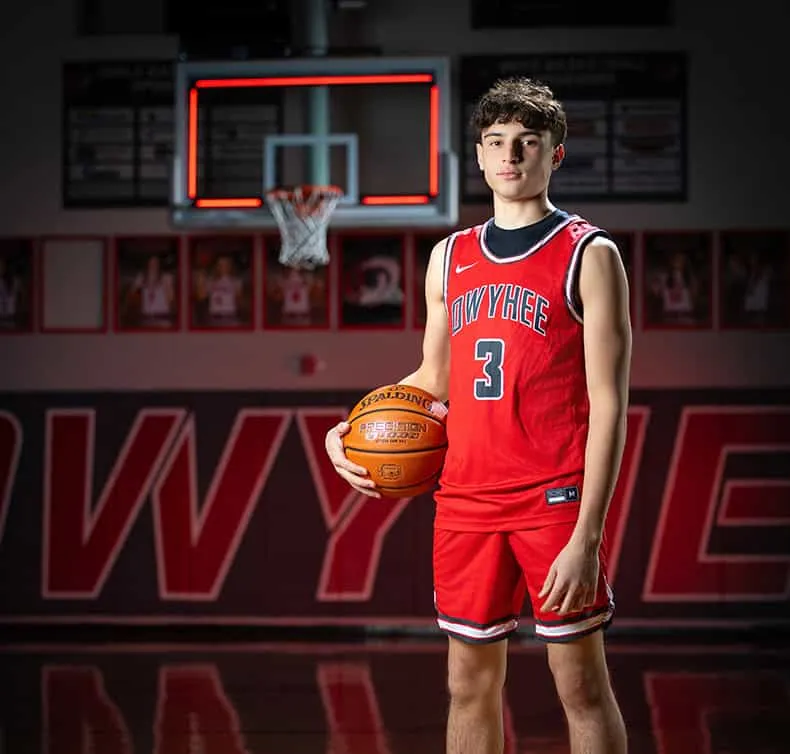 Owyhee high school basketball player in Nike red jersey number 3 holding Spalding basketball, indoor court with backboard and red neon lighting in background