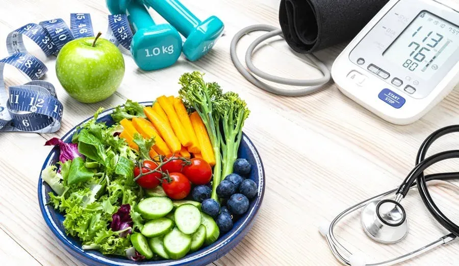 a health-themed setup, featuring a balanced and colorful salad bowl containing various fresh vegetables and fruits. Surrounding the bowl are health and fitness-related items, including:  A green apple. A blue measuring tape. A pair of blue dumbbells. A blood pressure monitor displaying readings. A stethoscope, indicating a medical or health check-up theme.