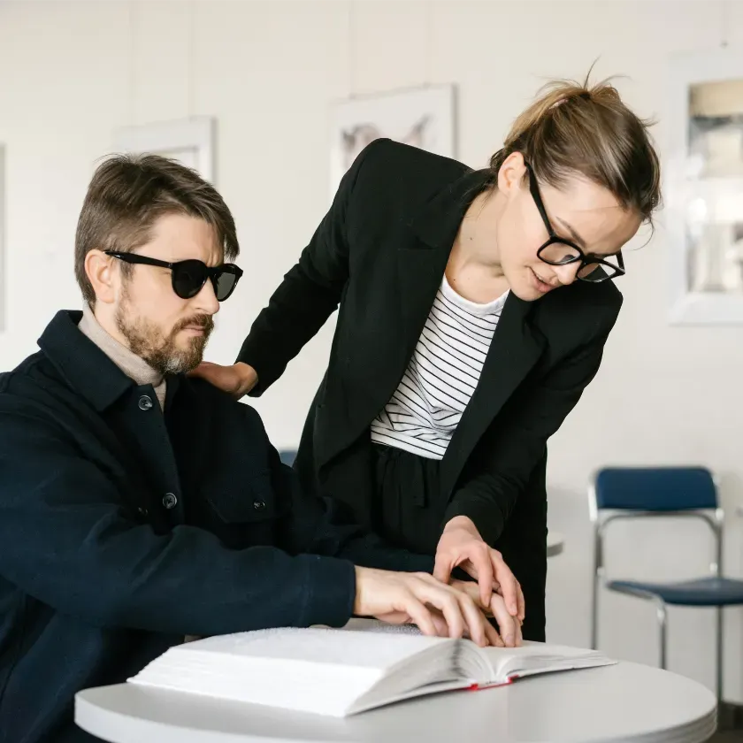 A woman wearing glasses and a black blazer assists a blind man who is reading Braille. The man, wearing sunglasses and a dark jacket, focuses on the book. They are in a modern, professional setting with blue chairs in the background. The word 'VISION' is displayed at the top.