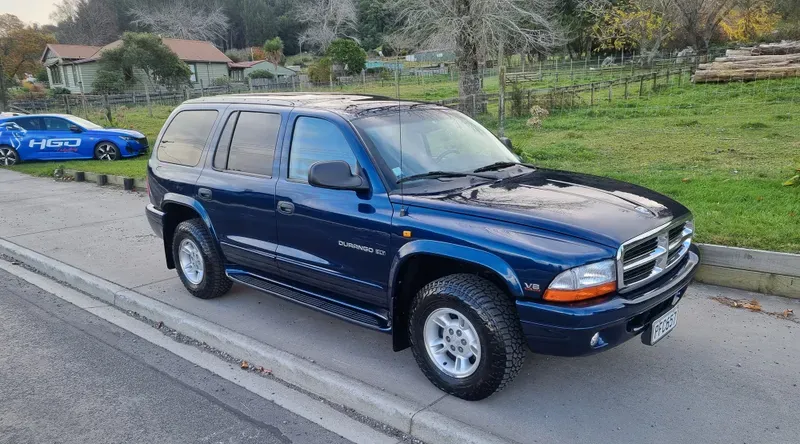 A dark blue Dodge Durango SUV is parked on the side of a suburban road. The vehicle features silver alloy wheels, tinted windows, and V8 badging. In the background, there are green fields, trees, and houses, with a blue car branded 