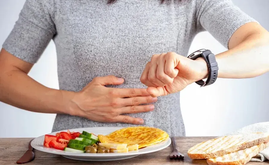 A woman sitting in front of a plate of food holding her belly while looking at her watch. She seems to be hungry and timing when she should start eating.