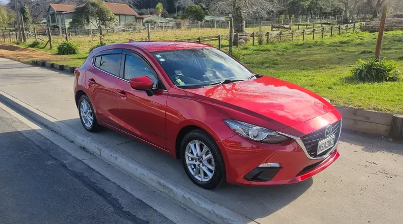 A red Mazda3 hatchback is parked on the side of a suburban road on a sunny day. The car features silver alloy wheels, a sleek design, and tinted windows. The background includes green fields, trees, and a few houses, with clear skies and bright sunlight reflecting off the vehicle's surface.