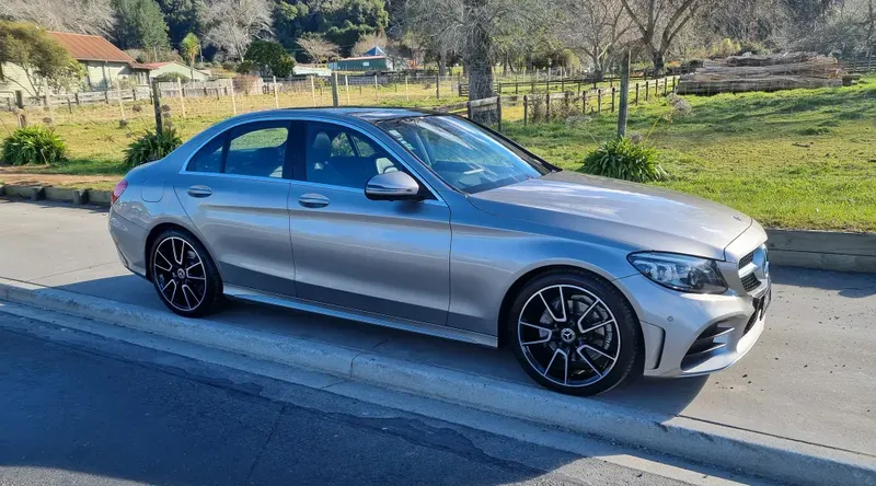 A silver Mercedes-Benz C-Class sedan is parked on the side of a suburban road on a sunny day. The car features black alloy wheels, a sleek design, and tinted windows. In the background, there are green fields, trees, and a few houses, with clear skies and a tranquil, rural setting.