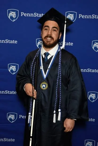 Young man standing in a blue graduation cap and gown holding his white cane. He is in front of a penn state banner.