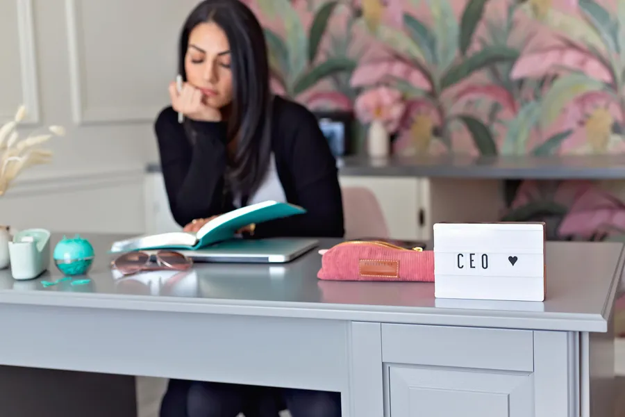 A woman sitting in front of desk table looking into a book holding a pen on her hand