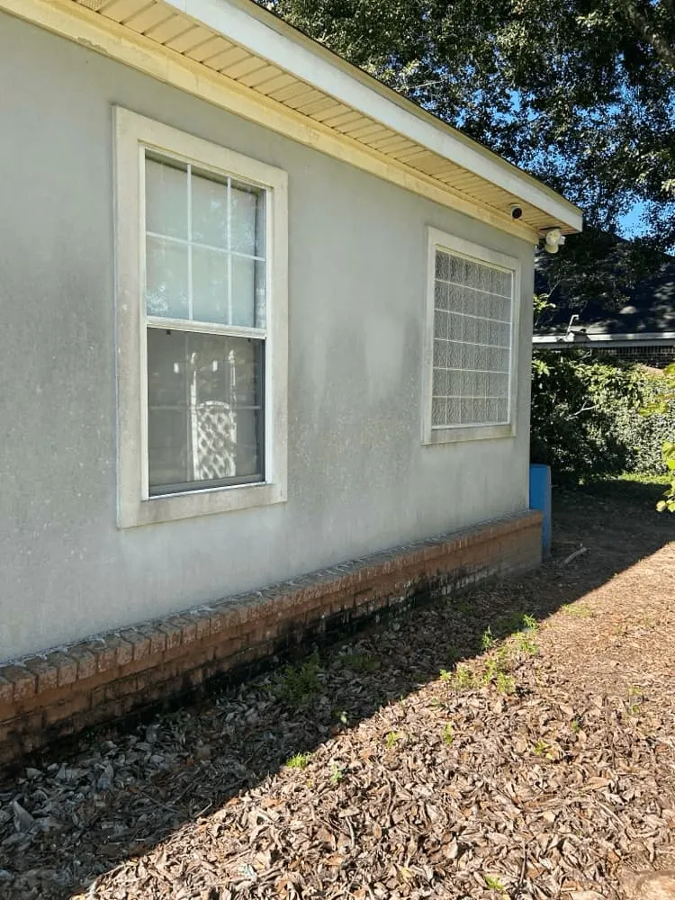 <alt=“Before stucco house soft wash in Dawes, Mobile County, Alabama showing dirt buildup on exterior walls and discoloration along brick foundation.”>