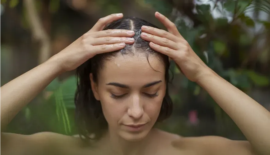 A close-up of a woman’s hands performing a hair and scalp massage using castor oil, with a serene expression, surrounded by nature and plants.