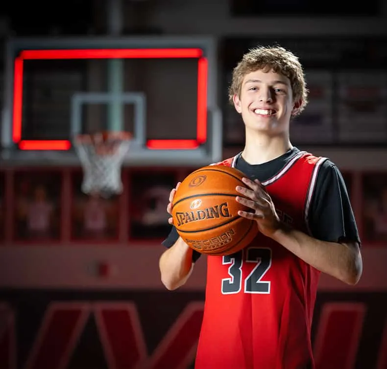 Owyhee High school basketball player number 32 holding Spalding basketball, smiling at camera in red jersey, basketball court and backboard with red lighting in background