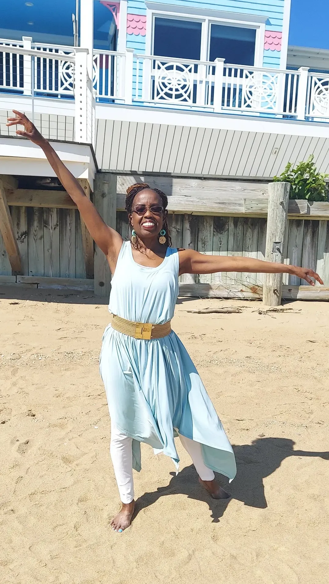 Coach Judy Handwerker stands barefoot on a sandy beach in front of colorful coastal homes, striking a graceful dance-inspired pose with one arm extended overhead and the other out to the side. Wearing a flowing light blue dress, white leggings, and a gold belt, she smiles confidently while expressing movement, balance, vitality, and joyful living. The beach setting reinforces themes of wellness, freedom, active aging, and holistic wellbeing.