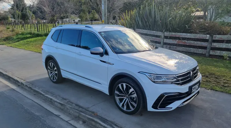 A white Volkswagen Tiguan SUV is parked on the side of a suburban road on a clear day. The vehicle features large alloy wheels, sleek lines, and tinted windows. The background includes a rural setting with green fields, trees, and a wooden fence, under partly cloudy skies.