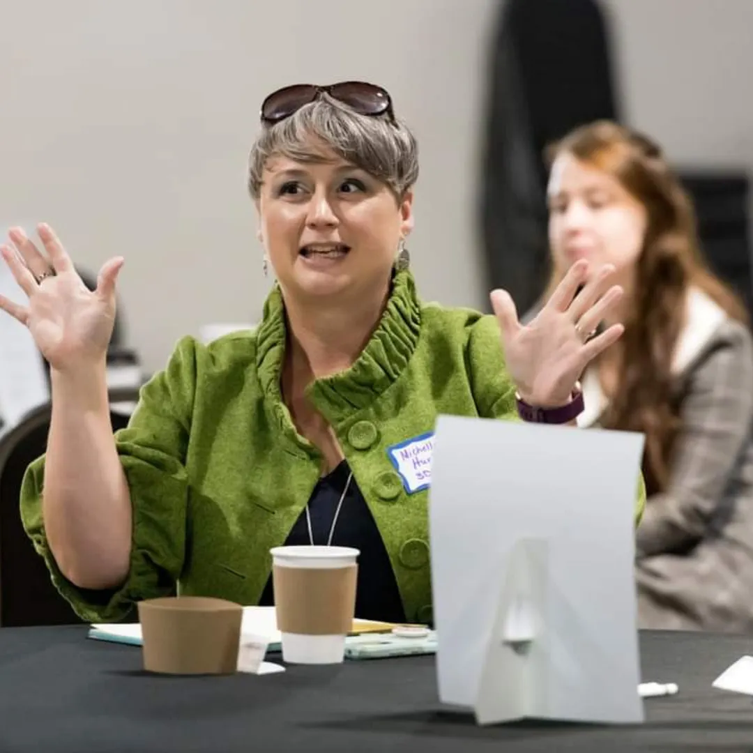 Director-Founder, Michelle Hurlburt sitting at a table with a black tablecloth. There is a coffee cup and a piece of notepaper in front of her. She is wearing a green jacket, her elbows are resting on the table with her hands up in their air as she is speaking. A pair of sunglasses sit on top of her head.