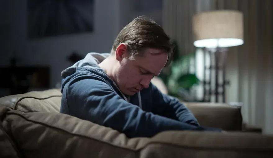 A man showing symptoms of vitamin D deficiency, conveying fatigue, depression, hair loss, and muscle pain in a dimly lit living room.