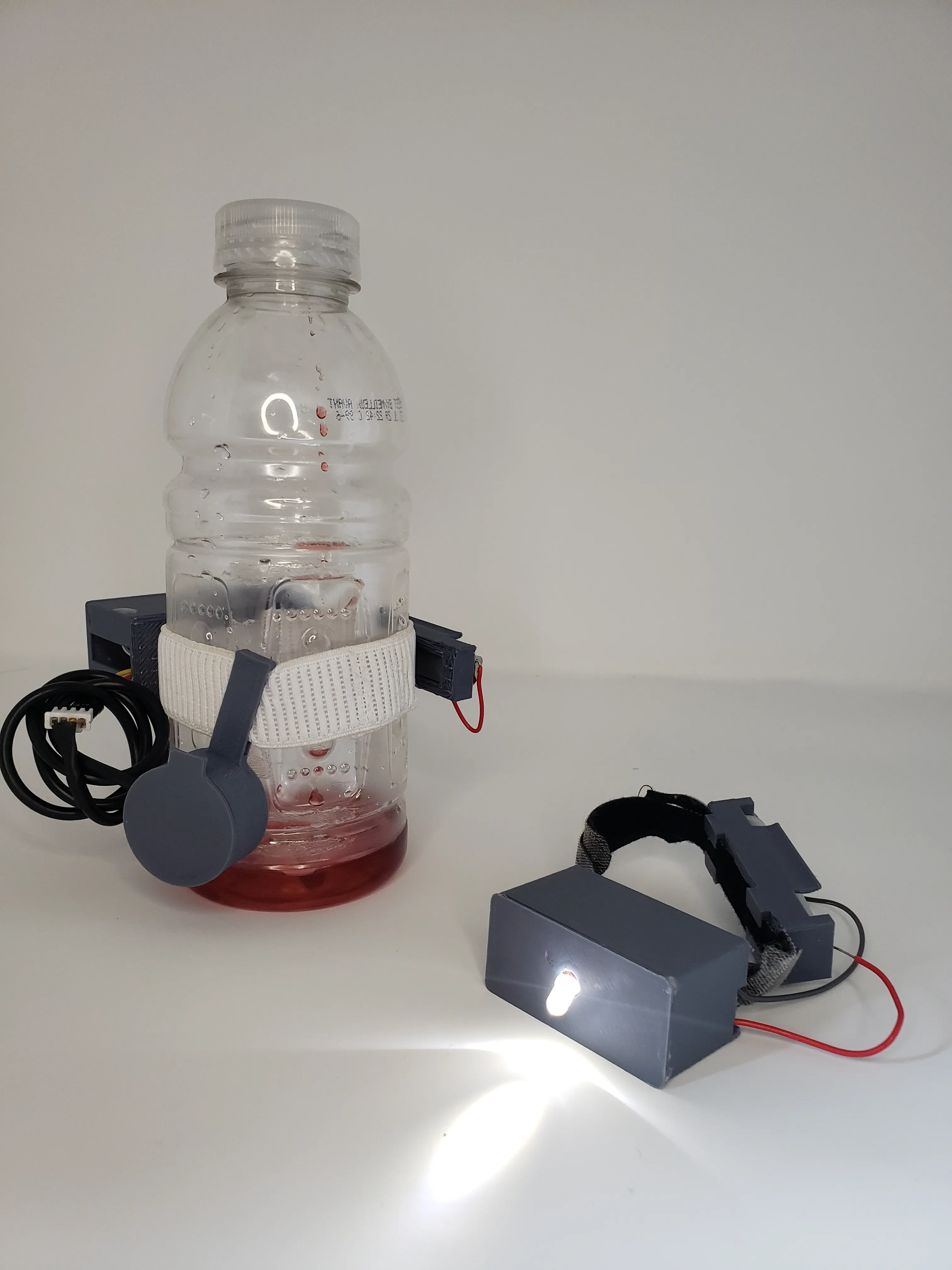 bottle filled with liquid lower than the non contact fluid sensor that is held onto the bottle with an elastic. Beside the bottle is a wrist band with an led on. 