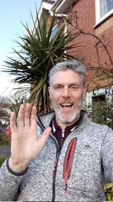 man waving at camera with house and palm tree behind