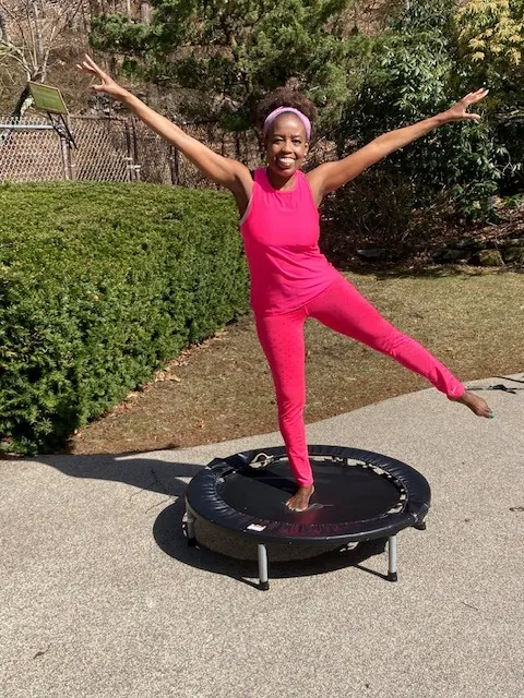 Coach Judy Handwerker demonstrates balance, strength, and joyful movement while standing on a mini fitness trampoline outdoors. Wearing a bright pink athletic outfit and matching headband, she balances on one leg with arms extended in a star-like pose. The sunny garden setting highlights themes of active aging, functional fitness, vitality, wellness, and confidence for women over 40.