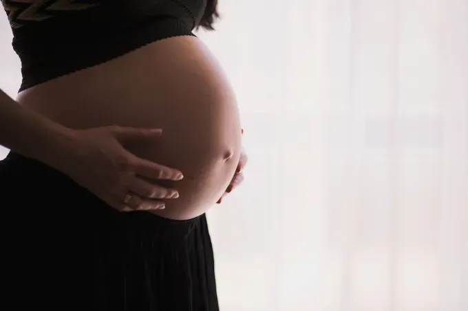 Pregnant woman cradling her belly with both hands, standing and smiling warmly.