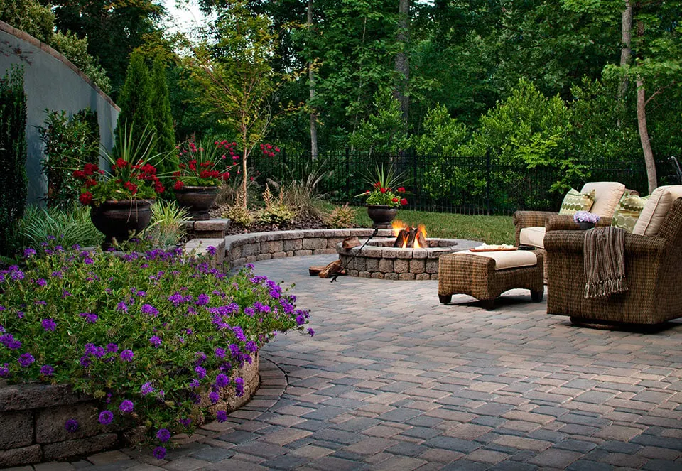 Beautiful alternating brick paver patio with recliner set, around a fire pit, and surrounded by red and purple flowers. 