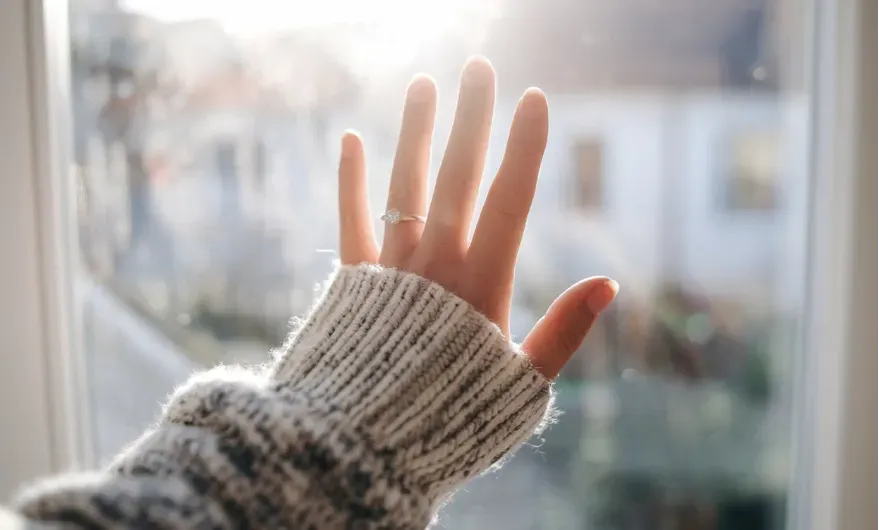 A woman's hand pressed against a sunlit window and half of the woman back of body is showing. The person is wearing a cozy, textured sweater, and a ring is visible on their ring finger. The light filtering through the window creates a soft, warm glow, casting a serene ambiance. In the background, an out-of-focus view of buildings or houses can be seen, suggesting an urban or suburban setting. The overall mood conveys a quiet, reflective moment, perhaps symbolizing longing or contemplation.