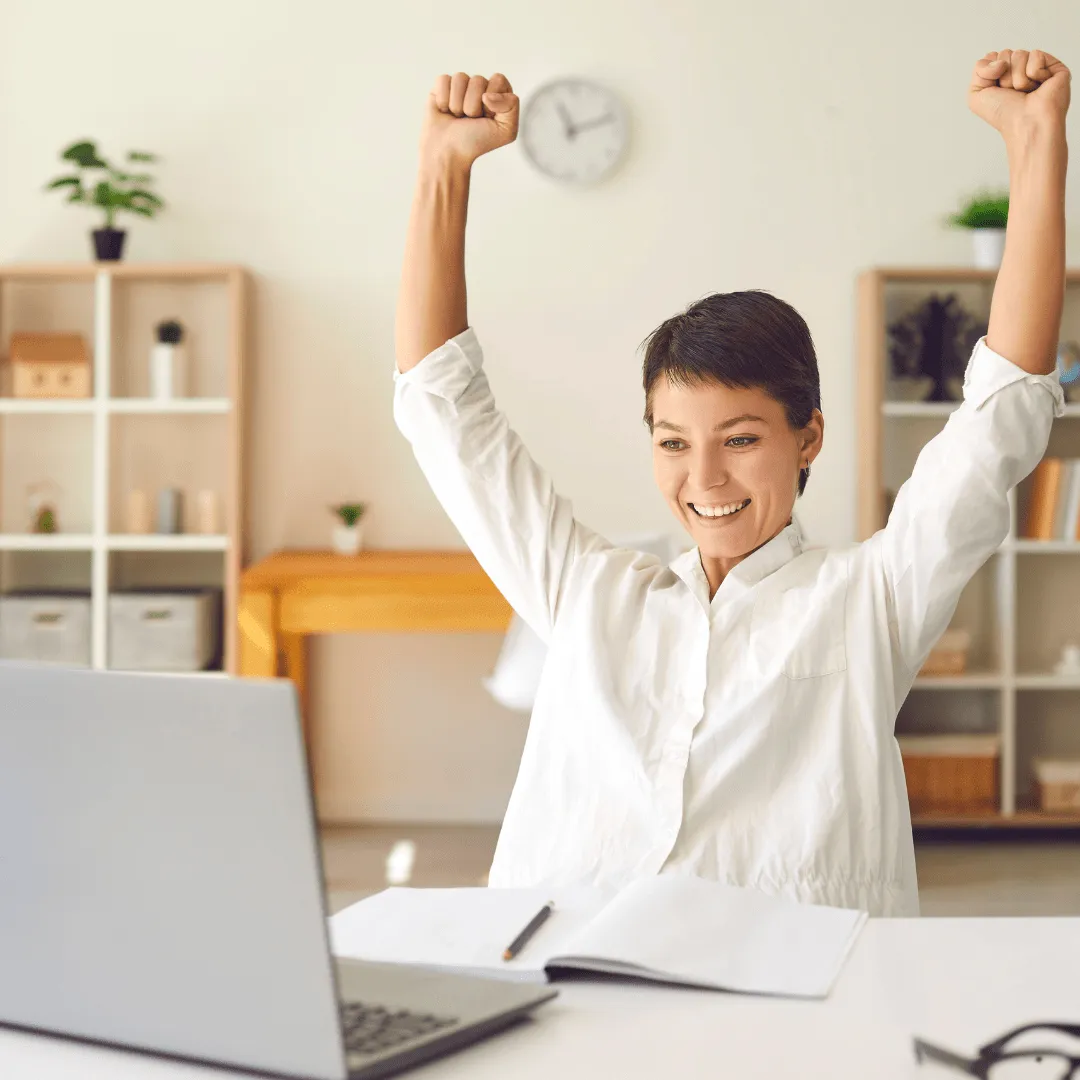 A happy woman sitting in front of her laptop with her arms raised. 