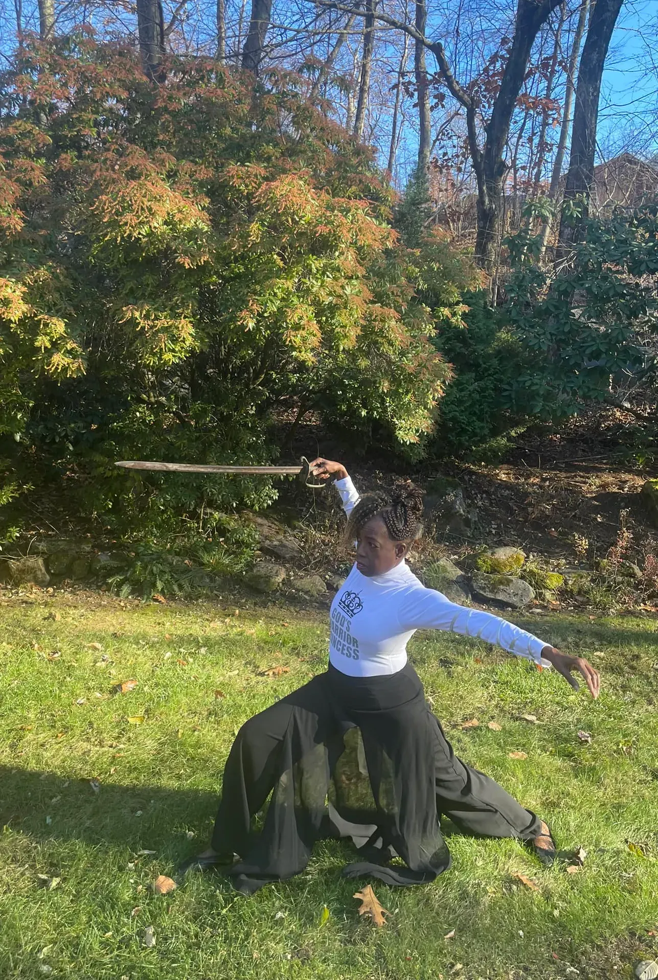 Coach Judy Handwerker performs a warrior-inspired movement pose outdoors, holding a sword overhead while kneeling in a graceful lunge on a sunlit lawn. Wearing a white 