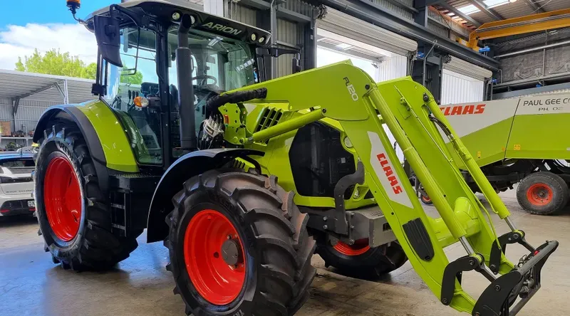 A bright green CLAAS Arion tractor is parked inside a large, well-lit garage or workshop. The tractor features large black tires with red rims, a front loader attachment labeled 