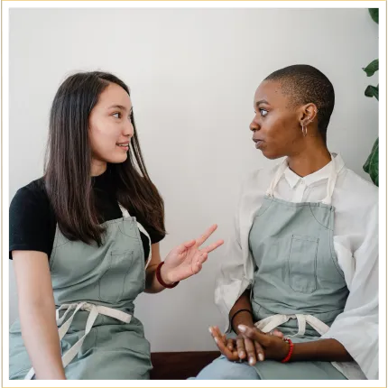 Two women wearing aprons one speaking the other listening