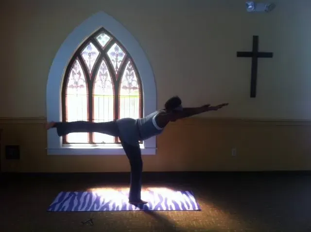 Coach Judy Handwerker demonstrates a balance and flexibility pose inside a church, standing on one leg with arms extended and the opposite leg raised behind her. Positioned on a yoga mat in front of a stained-glass window and cross, she showcases faith-based movement, strength, stability, and mindful wellness.